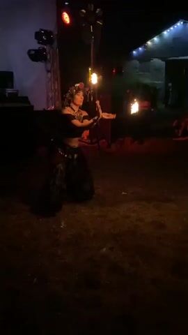 A person is performing with fire in a grassy outdoor area at night. The person is wearing loose-fitting pants with a patterned design and a dark, cropped top. They have a flower crown on their head and are holding two burning poi. The poi are creating trails of light as the person spins them. In the background, there are stage lights, a tent structure with lights, and other people who are not clearly visible. The ground is covered in dry grass and illuminated by the fire and stage lights. The lighting suggests a performance or festival setting.