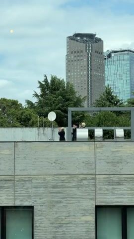 The video shows a wide shot of a rooftop with two individuals. In the background, there are tall buildings and trees. A faint yellow circle, possibly the sun or moon, is visible in the sky. On the rooftop, one person in dark clothing is interacting with a satellite dish. Another person, also in dark clothing with light-colored hair, is standing nearby. The rooftop appears to be made of concrete, with a metal railing and some greenery. The overall impression is an urban environment on a cloudy day.