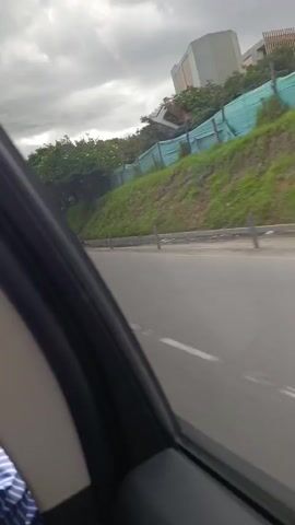 The video shows a view from inside a car moving along a highway on an overcast day. The camera looks out the passenger-side window, capturing passing scenery. The highway is separated from a grassy slope by a gray concrete barrier. On the slope, there's green netting and a fence. Trees dot the hillside. 

The car passes a scooter ridden by a person in red, and a yellow car. The camera briefly captures the interior of the vehicle, showing a blue jacket draped over a gray car seat and the interior trim of the car. The sky is gray and overcast.
