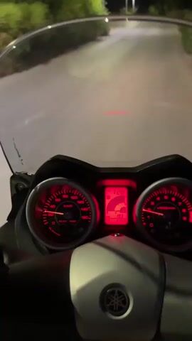 The video shows a first-person perspective of someone riding a motorcycle at night. The dashboard displays a speed of 40 km/h. The road is paved and lined with trees and streetlights. In the background, a body of water is visible with lights from a distant town on the shore, under a twilight sky.