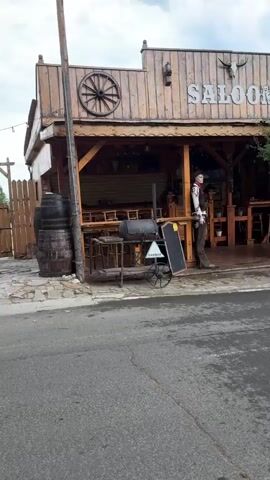 The video pans across the exterior of a themed establishment designed to resemble an Old West town. The front of the building features a prominent "SALOON" sign with a bull skull above it and a wagon wheel decoration. A mannequin dressed as a cowboy stands near the entrance, and a barbeque grill is positioned outside. To the right of the saloon, there is a smaller structure labeled "SHERIFF" with a jail cell visible inside, followed by a "POST" office and a building labeled "BANK". Wooden tables and chairs are set up on a covered patio area. The scene is set against a backdrop of trees and a cloudy sky. The street in front of the buildings appears to be paved.