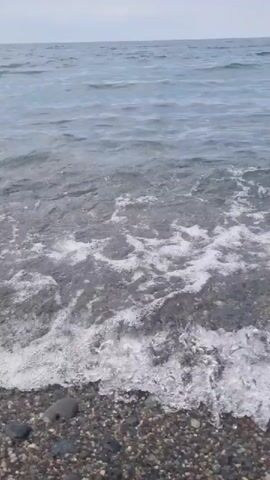 The video shows a close-up view of gentle waves washing over a rocky beach. The water is clear, and the pebbles on the seabed are visible. The sky is overcast, and the overall lighting is soft. The camera is positioned low, giving the impression of being at water level. There are no people or other objects visible in the frame. The sound is dominated by the lapping of waves.