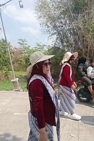 A group of women are walking outside. Some are wearing hats and sunglasses. One woman is wearing a blue shirt and a hijab. Another woman is wearing a maroon top and a white patterned skirt. There is a young girl in the foreground wearing a black shirt and white pants, with what appears to be money draped around her neck. In the background, there are trees and vegetation. The ground is paved. The sky is blue and there are some clouds.