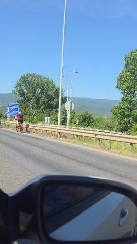 The video, filmed from inside a vehicle, presents a forward view of a multi-lane road on a clear, sunny day. In the lower foreground, the upper part of a vehicle's side mirror is visible, showing a blurred reflection. Two male cyclists, wearing helmets and athletic attire, are riding mountain bikes in separate lanes. The cyclist on the left wears a white top and black shorts, riding a dark-colored bike. The cyclist on the right wears a blue and black top with black shorts, riding a yellow-green bike. Both cyclists are approaching or stopped at a red traffic light. A large green tree stands prominent on the left side of the road, behind which a blue directional sign with white text is visible. Further in the background, green hills or mountains rise under a bright blue sky. Another cyclist is briefly visible ahead of the first two at the start of the video.