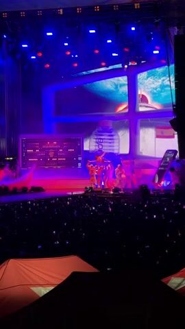 This video captures a live performance on a stage illuminated by blue and purple lights. In the foreground, a large crowd is visible, many holding up their phones. On stage, several performers are dressed in bright orange outfits, some in acrobatic poses. A large screen behind them displays images, including a giant water bottle and what appears to be a space-themed graphic. To the left of the stage, a monitor displays complex data, and a pinkish-orange dolphin-like sculpture is present. To the right, a Spanish flag is visible. The overall atmosphere suggests a concert or a theatrical production with elaborate staging and audience engagement.