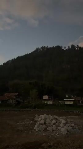 The video pans from left to right, showcasing a rural landscape at dusk. In the foreground, a pile of rocks rests on dark, disturbed earth. Beyond this, a small village is nestled at the base of a densely forested mountain. Several houses with rustic roofs are visible, some with lights on. The mountain itself is covered in dark green trees, and the sky above is a dusky blue with scattered clouds, tinged with the soft colors of the setting sun. The overall atmosphere is peaceful and quiet, evoking a sense of natural beauty and rural life.