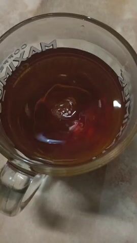 The video opens with a close-up shot of a clear mug filled with dark liquid, likely coffee or tea, showing ripples and bubbles on the surface. The mug has a handle and branding with the word "MAXIM" visible around the rim. The scene then shifts to a white plate filled with numerous small, pale dumplings, possibly pelmeni or pierogi, arranged in a cluster. A fork is partially visible in the upper right corner of the frame, suggesting the dumplings are about to be eaten. The background in both shots appears to be a speckled countertop.