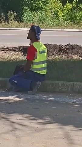A person wearing a blue cap, red shirt, blue work pants, and a yellow reflective vest is crouching next to a curb. The vest has text on the back. The person is looking to the left. Behind the person is a grassy area, then a road, and then trees and foliage. There are piles of dirt on the ground near the road. The foreground is a paved surface.
