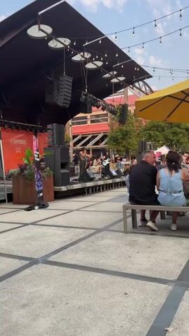 The video shows an outdoor performance with a band on a stage. The stage has speakers and musical equipment. A female singer is at the microphone in the center of the stage, wearing a patterned top and dark pants. A guitarist is to her right, also wearing dark clothing. Other band members are visible behind them. The audience is seated on benches in the foreground, facing the stage. There are string lights overhead, and trees and buildings are visible in the background. A yellow umbrella is to the right of the frame. The ground is paved, and the overall atmosphere appears to be casual and festive.