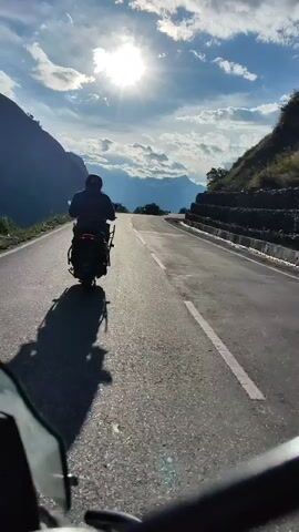 The video shows a person riding a scooter on a paved road. The road is curved and goes uphill, with mountains on either side. The sun is shining brightly in the sky, and there are clouds. The scooter rider is casting a long shadow on the road. The camera is positioned behind the scooter, suggesting it is being filmed from another vehicle or by a person riding behind the scooter. The view is from a first-person perspective of someone on a vehicle. The scene is a mountain road during daytime, with a clear sky and bright sunlight.