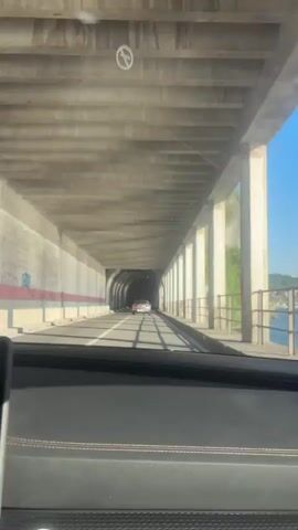 The video offers a first-person perspective from inside a car driving along a scenic lakeside road in Lierna, Italy. The car is moving under a concrete structure, resembling an open-sided tunnel or an overpass, which provides a partial canopy over the roadway.

At the beginning, the view is forward, showing the car's dashboard and the road ahead. Another vehicle is visible in the distance within the concrete structure. The overhead concrete beams and supporting pillars are prominent.

The camera then pans right, revealing a breathtaking view of the external environment. A clear, bright blue sky indicates sunny weather. Below the sky, a large body of water, consistent with a lake, stretches out, bordered by distant mountains. A black metal railing separates the road from the water. Concrete pillars, some marked with red and white safety stripes, line the right side of the road, with green foliage growing on the adjacent rock face or embankment. The sunlight is strong, suggesting it is midday or early afternoon. The scene depicts a picturesque drive along the shore of what is likely Lake Como, with no visible text or identifiable people.