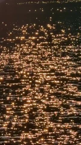 The video opens with a close-up of sun glinting off the surface of dark water. The camera then pulls back to reveal a lake surrounded by rocky shorelines and a dense forest of pine trees. In the background, a large mountain range is visible, with the sun setting behind one of the peaks, casting a golden glow across the water and the sky. The scene is serene and natural.