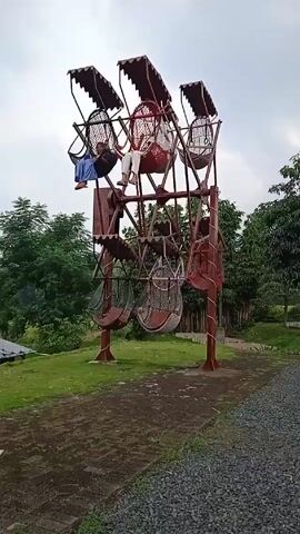 The video shows three people riding a stationary, non-motorized, rustic-style Ferris wheel on a cloudy day.  The wheel is constructed of metal painted in shades of maroon and brown, featuring six individual basket-like seats arranged around its perimeter. Each seat has a protective awning above. The riders are casually dressed, two are in the upper seats and one in a lower seat. The wheel is situated on a grassy field with surrounding trees. A path made of gravel runs in front of the structure. The sky is overcast.