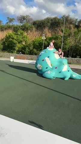 Two young girls are playing on a large, light blue climbing structure in an outdoor park. The structure is irregularly shaped, resembling an abstract animal or blob, and features various colorful handholds. One girl sits atop the highest point of the structure, while the other is positioned lower down, closer to the front. They appear to be enjoying their activity.

The ground beneath them is a green, soft-surface material, typical of playgrounds or recreational areas. In the background, a man can be seen riding a bicycle from left to right. Further back, there is a natural embankment or hill covered with trees and exposed earth, suggesting a landscape feature. Several streetlights are also visible, along with a white trash can and a wooden bench. The sky is partly cloudy but bright, casting long shadows on the green ground, indicating it is likely daytime, possibly late afternoon. The scene takes place in Taman, Russia.