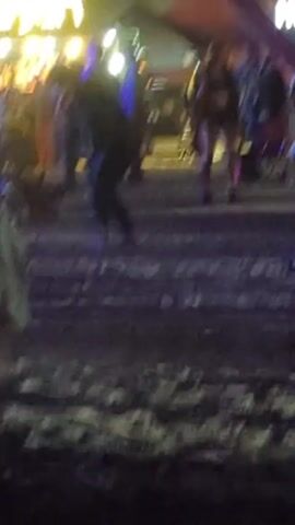 The video shows a night scene, possibly a party or gathering, with people moving around in the background. A light fixture with Hebrew writing illuminates a food stall or bar in the background. In the foreground, a tall object wrapped in cloth is visible. The ground appears uneven, possibly dirt or sand, and is illuminated with blue and purple lights. People are walking, standing, and interacting in the background, some moving towards the food stall. The overall scene appears lively and active, with background noise suggesting music or conversation.