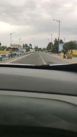 The video shows a view from inside a car as it drives down a highway. The car's dashboard is visible in the foreground, and water droplets are on the windshield, suggesting it might be raining or recently rained. The highway appears to be a multi-lane road with a median strip. There are streetlights lining the road, and trees and buildings are visible on either side. In the distance, other vehicles can be seen on the highway. There is a billboard on the right side of the road. The sky is overcast, indicating a cloudy or possibly stormy day. The overall mood of the video is mundane, depicting a typical car journey. No people are clearly visible. The location cues are consistent with a highway in a semi-urban or suburban area.