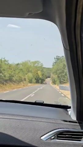 The video shows a view from inside a car, looking out the windshield. The car is driving on a paved road with white lines marking the lanes. There are trees and greenery on both sides of the road. In the distance, there are two cars driving ahead. The sky is visible above and appears to be clear. The interior of the car is visible in the foreground, including the dashboard and part of the car's A-pillar. The overall scene suggests a daytime drive on a country road.