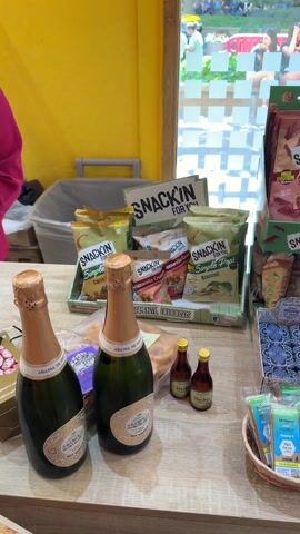 The video shows the interior of a small store or kiosk, looking out through a large window. In the foreground, there's a table laden with snacks including bags of "SNACKIN' for you" chips, bottles of what appears to be champagne, and small bottles of liquor. Additionally, there's a display of chewing gum and other small snack items.

Looking through the window, one can see an outdoor setting. A group of young women, mostly wearing shorts and t-shirts, is sitting on a low wall or ledge.  Beyond them, there's a crowd of people and greenery, suggesting a park or campus setting. The architecture in the background suggests an urban environment. A man walks through the store door, looking at his phone.