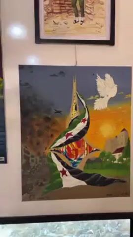 The video shows a close-up of a wall displaying a collection of framed artworks. The paintings vary in style and subject matter. One painting depicts a cityscape with a river, modern buildings, and futuristic elements. Another painting shows a scene of war and destruction on one side, transitioning to a peaceful landscape with a white dove on the other side, bisected by the Syrian flag. Above them are two more paintings, one depicting a person standing near a building and another with a group of people. A miniature version of Van Gogh's "Starry Night" is also visible among the paintings.