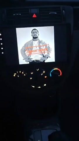 The video shows the dashboard of a car as it drives down an open road. The dashboard includes a radio playing music, a temperature gauge, and other controls, all illuminated. The view through the windshield shows a flat, empty road with a cloudy sky. The video seems to have been taken from the driver's perspective.