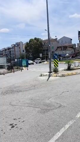 The video shows an outdoor scene on a sunny day with a blue sky and scattered clouds. In the foreground, there is a paved road with some dark stains. To the left and center, there are partially constructed buildings surrounded by fences. Further back, several yellow taxis are parked. A man in a dark shirt is walking between the taxis. In the background, there are more buildings and some debris piles. A traffic light and a pedestrian crossing are visible on the right side of the road. The overall impression is of an urban area undergoing construction, with taxis present, possibly indicating a taxi stand or waiting area.