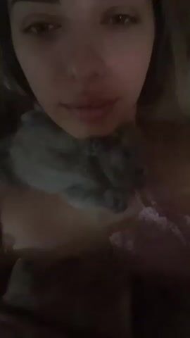 The video shows a close-up, selfie-style shot of a person's face and upper chest. The person has dark hair and eyebrows, and their eyes are open, looking towards the camera. They are wearing a pink lace-trimmed garment. A small gray animal, possibly a hamster, is resting on the person's chest, near their neck. The lighting is dim, with the person's face illuminated. The background is dark and indistinct. The video is very short, lasting only a few seconds.
