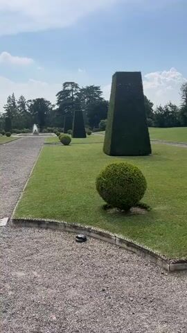 This video showcases a meticulously manicured garden with a distinct formal style. The primary subjects are large, conical and spherical topiaries, expertly shaped from dark green hedges. These sculptures are strategically placed across a vast expanse of vibrant green lawn.

In the foreground, a rounded topiary sits close to the camera, its dense foliage a rich green. To the left, a gravel path runs along the edge of the lawn, bordered by a decorative rope-like edging. A small, dark, circular object, possibly a garden maintenance robot, is visible on the gravel.

Further into the scene, three tall, tapering cone-shaped topiaries stand in a row, creating a strong vertical element against the horizontal sweep of the lawn. In the distance, a fountain can be seen on the left, hinting at a larger estate or park. The background is filled with mature trees, some with characteristic Italian cypress shapes, and a bright blue sky with scattered white clouds, suggesting fair weather.

The overall impression is one of order, grandeur, and serenity. The sunlight indicates it is daytime, likely late morning or afternoon, with clear skies. The setting evokes a sense of a well-established, possibly historic, garden in Varese, Italy. No people are visible, and no text is present in the media. The activity taking place is simply the presentation of the garden itself, highlighting its structured beauty and the impressive horticultural skill involved in its upkeep.