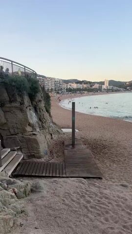 This is a vertical, low-angle, static shot of a beach in the late afternoon or early evening. The sky is a clear, pale blue with a hint of pink near the horizon. The sand is a light brown and textured with footprints and ripples. In the foreground, a wooden walkway leads from the sand towards a rocky cliff face on the left. A simple outdoor shower structure stands on the walkway. The midground shows a wide expanse of sandy beach dotted with people. Some are sitting on towels, others are walking or standing. A few people are in the gentle waves of the calm, light blue sea. The background features a long, curving coastline lined with multi-story buildings, suggesting a town or resort. The buildings are white and cream-colored, with many balconies and windows. Further in the distance, hills rise behind the town, and a tall white building stands out against the landscape. The overall atmosphere is peaceful and relaxed, typical of a beach scene at the end of the day.