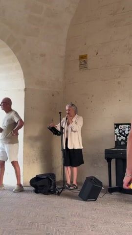 A woman stands under an arched stone structure, singing into a microphone positioned on a stand in front of her. She wears a light-colored jacket over a dark top and shorts, with sandals. To her right, a black keyboard sits on a stand. A black speaker is placed on the ground in front of the woman, and another black bag rests behind her. A person walks past in the background on the left. The floor is paved with bricks.