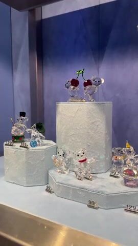 The video showcases a display of Swarovski crystal figurines in a retail setting. The figurines, primarily clear crystal with subtle colored accents, depict teddy bears and other festive motifs like cherries and a snowman. They are arranged on tiered light blue platforms with price tags visible. The background is a textured blue wall, and the display is enclosed within a glass case. No people are visible in the shot. The camera pans slightly, offering different angles of the display.