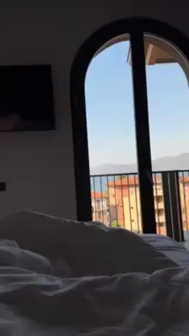 The scene is an interior shot of a bedroom with a rumpled white bed in the foreground. In the background, a large arched window or door leads to a balcony. The view from the balcony includes buildings with red roofs, a body of water, and distant mountains under a clear blue sky. A large black television screen is mounted on the wall to the left of the window. The room appears dimly lit, casting shadows on the bed and the wall. The camera slowly pans across the room.