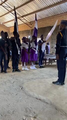 The video shows a group of people standing in what appears to be an indoor space with a dirt floor and a corrugated metal roof.  On the left side of the frame, several individuals in dark uniforms, some holding what look like rifles, are standing at attention.  Two individuals, one in a dark uniform and the other in a white shirt and dark pants, are holding large flags.  In the center, two young females wearing purple skirts and white tops are standing in formation, also holding flags.  To their right, a person in a dark uniform is holding a white cloth.  On the far right, a person in a dark uniform with yellow accents is standing facing the group.  The overall scene suggests a ceremonial event, possibly a flag presentation or a similar solemn occasion. The environment is rustic and unadorned.
