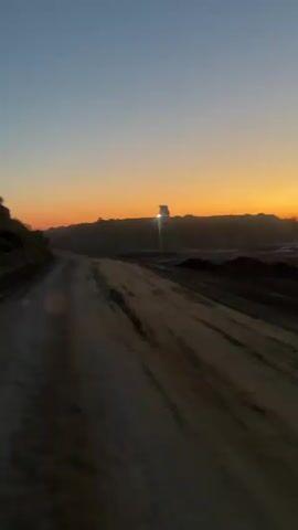 The video captures a dirt road during sunset. A vehicle with its headlights on is visible in the distance, silhouetted against the orange and blue sky. The road is dusty and appears to be in a rural or industrial area. The sun is low on the horizon, casting a warm glow.