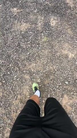 The video shows a first-person perspective of someone walking on a rocky, arid landscape. The person is wearing black shorts, white socks, and bright green running shoes. They are walking towards a rugged, mountainous terrain under a cloudy sky. The ground is covered in small grey and brown rocks.