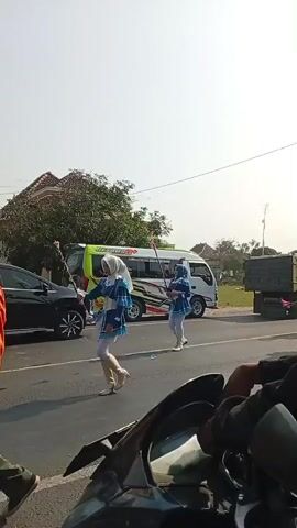 The video shows a street scene under a bright, clear sky. In the foreground, the handlebars and instrument panel of a motorcycle are visible, suggesting the video is being filmed from a rider's perspective.

On the street, a small parade or procession is taking place. Several individuals, primarily young women, are dressed in matching blue and white marching band uniforms. They are wearing headscarves, also in matching colors. Some are carrying what appear to be batons, and they are walking in a line along the side of the road. Their movements suggest they are part of a marching drill or performance.

Behind the marchers, a white and yellow minibus with colorful graphics is parked or driving slowly. Further down the road, a truck is visible, possibly part of the procession or a vehicle belonging to the group. To the left of the procession, a black car is parked, and beyond that, trees and buildings can be seen, indicating a semi-urban or rural setting. The overall atmosphere appears to be sunny and casual, likely during a daytime event.