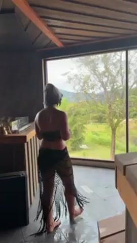 The video shows a person standing in a room, facing a large window that overlooks a lush green landscape with mountains in the background. The person is wearing a black top, a black fringed skirt, and a hat or towel wrapped around their head. They hold a round, flat drum with a handle and appear to be preparing to play it. The room has a wooden ceiling and tiled floor. A wooden bar and other furniture are visible in the background.