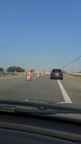 The video captures a first-person perspective from inside a moving vehicle on a highway during daytime. The sky is clear and blue, indicating sunny weather. The road ahead is asphalt, marked with white lines. Numerous red and white striped pylons with orange tops are placed on the road, suggesting a construction zone or lane closure.

Ahead of the viewer's vehicle, a dark-colored SUV is driving in the right lane, following the path marked by the pylons. Further in the distance, a white van and another dark car are also visible. On the left side of the highway, green trees and vegetation can be seen, while a metal fence-like structure runs along the right side. The overall scene depicts typical highway travel through a marked road work area. No specific text is legible within the video frame. The location is Zavoy, Bulgaria.