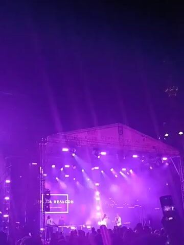 The video captures a lively outdoor concert or event at night. The stage is illuminated with bright purple and white lights, creating a dynamic atmosphere. A large screen displays images of a woman, likely the performer, singing into a microphone. The screen shows different shots of her, sometimes in close-up, wearing a white outfit. Behind the screen, several people are visible on stage, possibly musicians or crew members, working with equipment. The foreground is filled with the silhouette of a crowd of people, many holding up their phones to record the performance. The overall impression is one of an energetic and engaging live music experience.