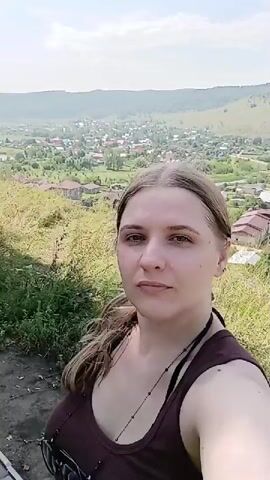 A woman with light brown hair pulled back in a ponytail and wearing a dark brown tank top is taking a selfie outdoors. She is looking at the camera with a neutral expression. In the background, there is a scenic view of a town nestled in a valley, with rolling hills and trees under a bright, sunny sky. To the left of the woman, two other people are visible sitting on a wooden bench. One is a man wearing a striped shirt and shorts, and the other is a woman wearing a light blue t-shirt. They appear to be engaged in conversation. The overall setting suggests a casual outing in a picturesque natural landscape.