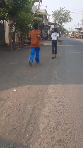 Two young boys are running away from the camera down a paved road. The boy in front is wearing an orange shirt and blue pants. The boy behind him is wearing a white shirt with a black stripe down the side and black pants. There are buildings on the left side of the road, and trees and utility poles in the background. The sun is shining, casting shadows.