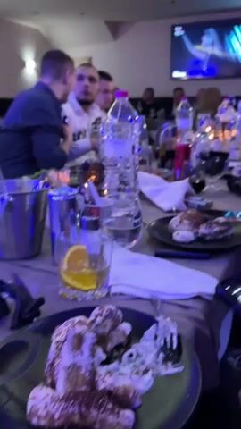 The video shows a group of men sitting around a long table, appearing to be in a restaurant or banquet hall. The table is laden with food plates, glasses, water bottles, and other items.  There are multiple men, mostly young, seated around the table, some wearing casual clothing. One man raises his glass. In the background, a television screen displays a music video. The overall atmosphere seems social and celebratory.