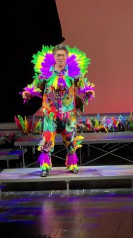 A male performer stands center stage in a brightly colored, elaborate costume adorned with large, vibrant feathers in neon green, purple, and yellow. The costume features a tropical print shirt and pants, with matching feather accents on his collar, wrists, and ankles. He wears dark shoes. The performer has his mouth open, appearing to be singing or speaking, with his arms extended slightly. Behind him, on tiered risers, are rows of similar colorful feathered props. The stage is dimly lit, with a dark backdrop on the left and a pinkish-red gradient on the right. The floor in front of the stage is dark and appears to be reflecting stage lighting in purple and green hues.