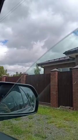 The video shows a view from inside a car, looking out the passenger side window. A brown sedan is visible in the reflection of the side mirror. The car appears to be moving along a gravel road. On the right side of the road, there is a tall, dark brown metal fence with brick pillars. Beyond the fence, a house with a dark roof is partially visible. The sky is partly cloudy. Further down the road, a lighter colored metal fence appears, followed by a green wooden fence. Lush green vegetation and trees are visible along the side of the road and behind the fences. The overall environment suggests a rural or suburban setting.