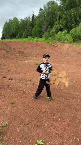 A young boy in a long-sleeved shirt, black pants, and a baseball cap is standing on a red dirt field, holding a long stick. He raises the stick as if to swing it, then lowers it and smiles at the camera. In the background, there are green trees and a cloudy sky.