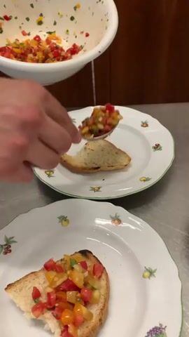 A person's hands are shown preparing bruschetta. A white bowl filled with chopped tomatoes, peppers, and herbs is held above two white plates, each with a slice of toasted bread. A spoon is used to transfer the tomato mixture from the bowl onto the bread. One plate has a serving already placed, while the other is being served. The background appears to be a kitchen or dining area with wooden cabinetry and a metallic countertop.
