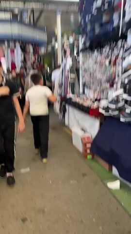 The video pans through a busy market, likely an indoor bazaar. The camera follows a young boy in a white t-shirt and black pants walking through a crowded aisle. To the left, a woman in a green patterned outfit and a red headscarf walks towards the camera. Behind her, other shoppers are visible, some wearing hijabs. To the right of the aisle, market stalls are packed with merchandise, including clothing, accessories like hair clips and jewelry, and shoes. Fabrics and garments hang from displays. The ground is concrete. The lighting appears to be a mix of natural light from overhead and artificial lights from the stalls. The overall atmosphere is one of commerce and everyday activity.