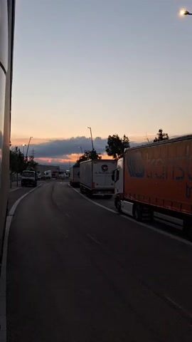 The video shows a line of parked trucks on a paved road. The sky in the background is a clear gradient from light blue at the top to a warm orange and pink near the horizon, with dark clouds scattered across it. The sun appears to be setting, casting a soft light over the scene. Streetlights are visible along the roadside, and trees can be seen in the distance. The trucks are of various sizes and colors, with some having company logos or text on their trailers. The camera is positioned from the perspective of someone or something alongside the road, looking down the lane where the trucks are parked. The overall atmosphere is calm and serene, with a focus on the industrial setting contrasted with the natural beauty of the sunset.