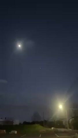 The video shows a night sky with a bright moon and a few visible stars. The camera pans slightly, focusing on one particularly bright star. In the foreground, there is a parking lot with concrete barriers, a street lamp, and some foliage. The overall atmosphere is dark and still.