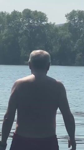 A man is standing in shallow water at the edge of a lake. He is facing away from the camera, with his back to the viewer. He is wearing red swim trunks. The water is calm, and there are trees on the opposite shore. The sun is shining, creating ripples on the surface of the water. The man's skin is visible, and he has short, graying hair. He appears to be enjoying the water.