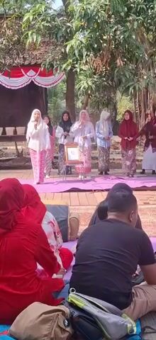 The video shows an outdoor event with a group of women on a raised wooden stage, some wearing hijabs and traditional attire. One woman in a white top and patterned skirt stands at the far left of the stage, holding a microphone and speaking. Next to her, another woman in a black hijab holds papers. In the center of the stage, a woman in a white hijab and patterned skirt stands behind a framed picture on a stand. Other women in similar attire stand to her right. The stage is decorated with red and white fabric at the back. In the foreground, an audience sits on the ground, facing the stage. Several people are wearing red clothing, and others are in casual wear. The environment appears to be a park or garden, with trees and foliage visible in the background. The lighting suggests it is daytime.
